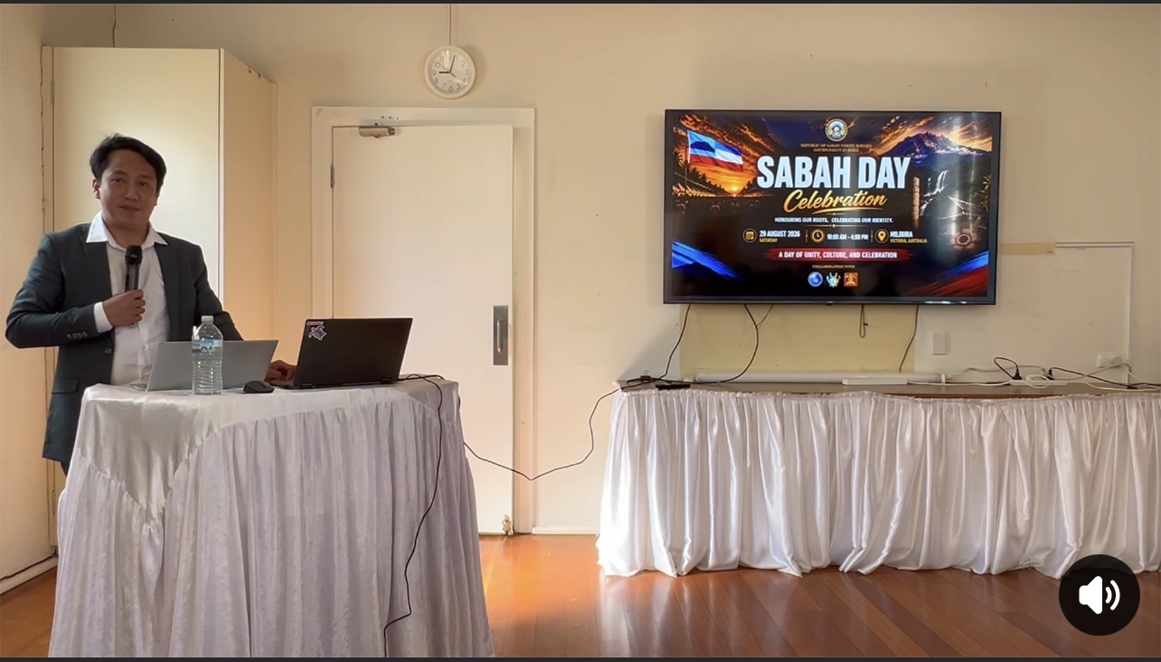 Screenshot of a speaker holding a microphone beside a Sabah Day celebration display